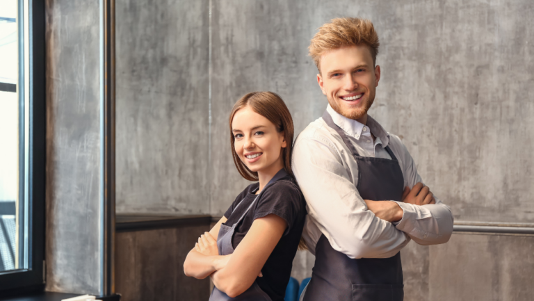 A man and woman in aprons standing back-to-back with arms crossed, smiling confidently in a modern, industrial-style interior.